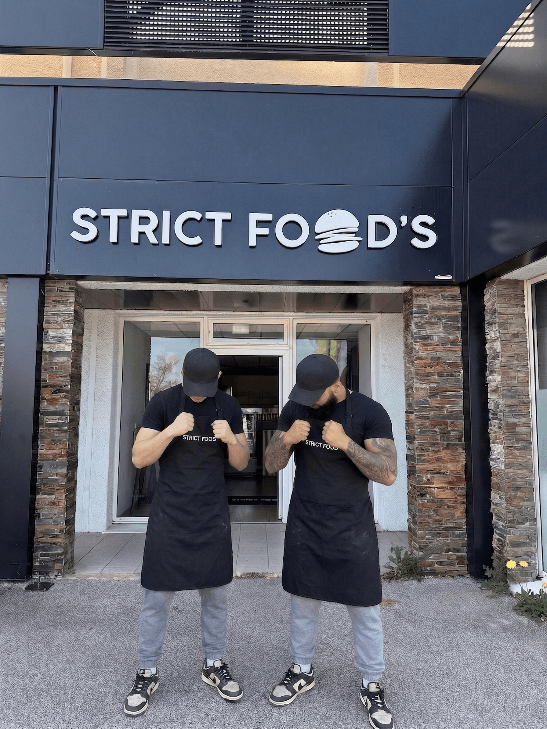 Les deux fondateurs de StrictFood devant la devanture du restaurant, casquettes baissees, tabliers noirs, poings leves